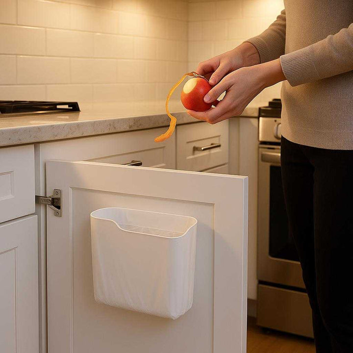 Affaldsposeholder til vægmontering - Moderne og pladsbesparende løsning til køkkenet. A sleek, modern kitchen with white cabinetry and stainless-steel appliances. The affaldsposeholder is mounted discreetly inside a cabinet door, lined with soft under-cabinet lighting casting gentle shadows. The kitchen glows with a warm, inviting ambiance. A person is casually peeling an apple over the holder, effortlessly disposing of the waste, showing its ease and practicality.