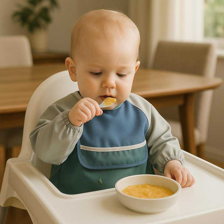 Anti revers bib i polyester, vandafvisende, dækker hele overkroppen, i farverne sortgrøn, beige, lyserød og blå: A realistic family dining room setting where the bib is properly grounded on a highchair, being actively used by a baby during mealtime.