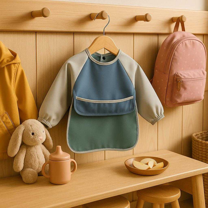Anti revers bib i polyester, vandafvisende, dækker hele overkroppen, i farverne sortgrøn, beige, lyserød og blå: A different realistic daycare setting where the bib is securely positioned on a coat rack among children’s belongings.