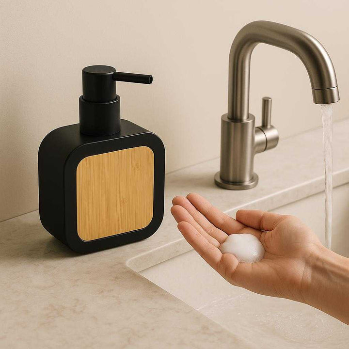 Bambus Sæbepumpe - Elegant og Funktionel Design til Badeværelset med Bambus Dekoration. In a minimalist bathroom setting, the elegant black soap dispenser with bamboo decoration is placed beside a running faucet, dispensing soap into a person's hand over a marble sink.