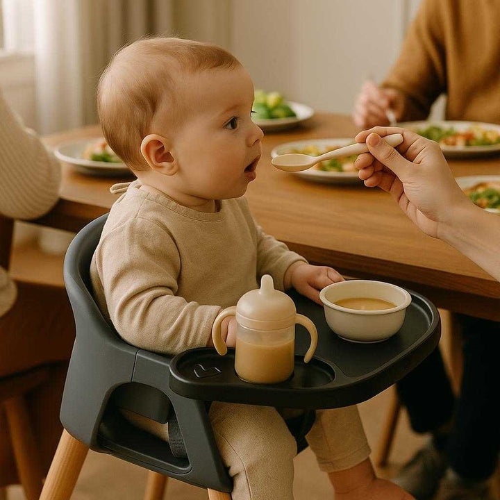 Barnevognsbakke i sort plast - Praktisk og moderne design med nem rengøring. The product is elegantly installed on a stylish high chair indoors, showcasing its practicality by holding a baby’s feeding essentials during a lively family lunch.