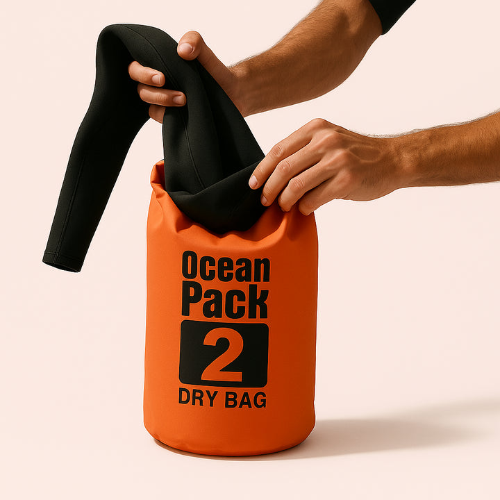 Enhanced Bådtaske: Vandtæt og Slidstærk Drybag – Ideel til Outdoor & Vandsport – Tilgængelig i Orange, Sort, Himmelblå, Grøn og Størrelser 2L, 3L, 15L: A realistic beach setting where the orange drybag is properly grounded on the sandy shore, being actively used by a surfer who is placing their wet suit inside it, with natural lighting casting realistic shadows and showing clear contact points where the bag touches the sand.