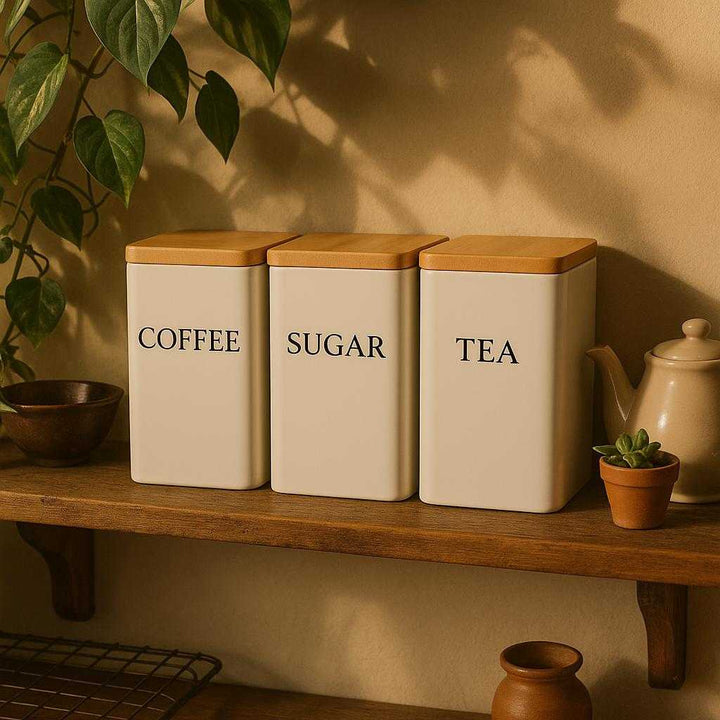 Beholdere til kaffe, te og sukker med hvidt og sort design – Høj Kvalitet og Trælåg. In a cozy kitchen setting, the elegant coffee, tea, and sugar holders with wooden lids are displayed on a rustic wooden shelf, surrounded by plants and vintage kitchenware, casting soft shadows.