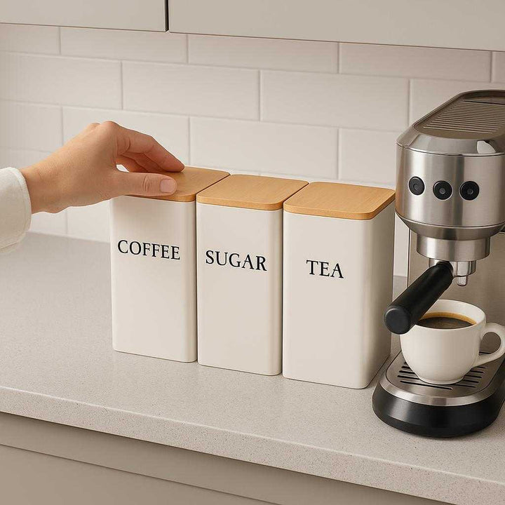 Beholdere til kaffe, te og sukker med hvidt og sort design – Høj Kvalitet og Trælåg. The high-quality white and black designed coffee, tea, and sugar holders are neatly arranged on a modern kitchen countertop, with a hand reaching to open the sugar holder for a fresh cup of coffee being prepared beside a stylish espresso machine.