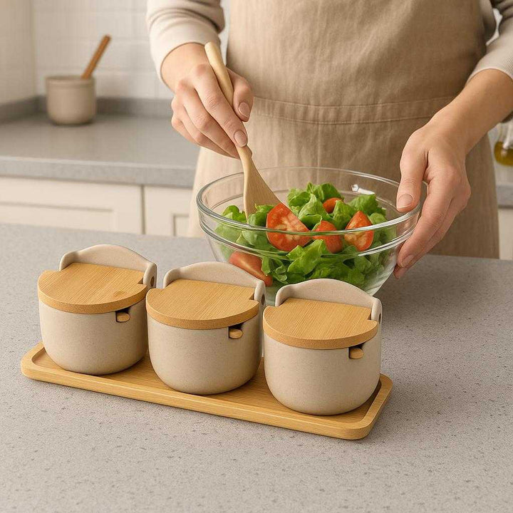 Beholdere til Krydderier - Moderne Sæt med 3 Keramiske Beholdere og Bambuslåg - Beige og Lys Brun. The beige and light brown ceramic spice containers, with bamboo lids slightly ajar, are neatly arranged on a kitchen counter, beside a person preparing a salad with various spices within reach.