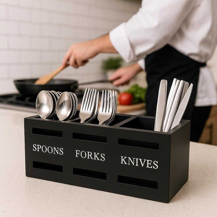 Bestikholder i Sort Træ med 3 Rum – Professionel Organisering til Køkkenet. The sleek black wooden cutlery holder sits organized on a bustling kitchen countertop, filled with stainless steel spoons, forks, and knives as a chef prepares a meal nearby.