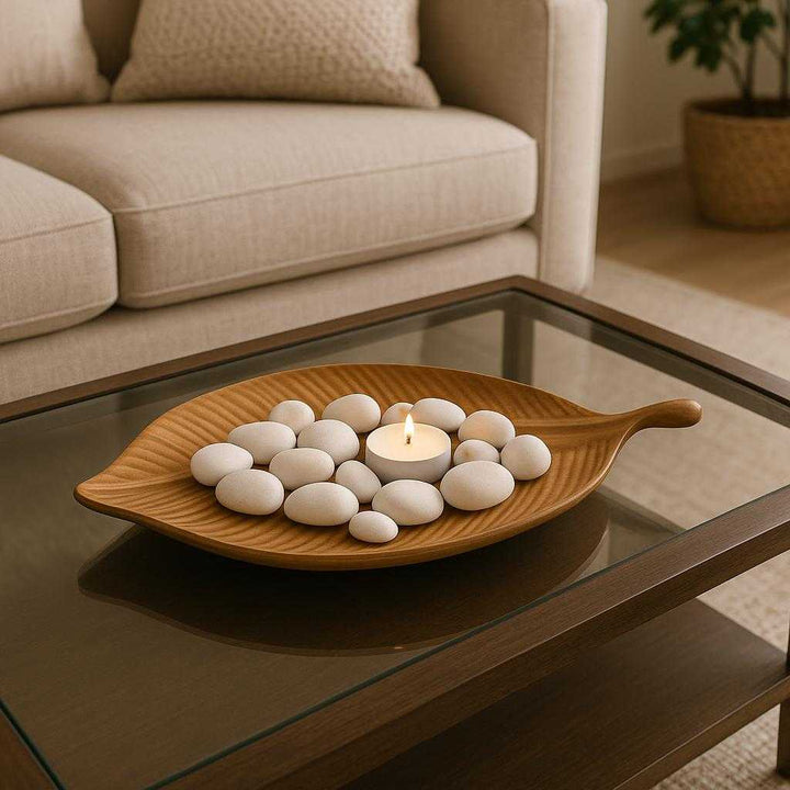 Bladformet træfad i naturtræ - Elegant og Funktionel Dekoration til Moderne Hjem. Placed on a polished living room coffee table, the leaf-shaped tray elegantly holds decorative white stones and a small candle, complementing a modern home setting with a cozy atmosphere.