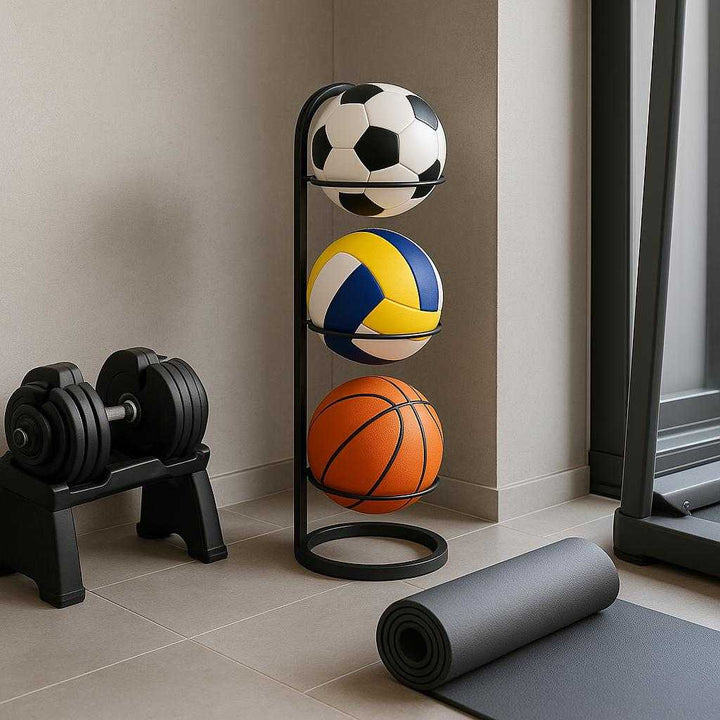 Boldholder til 3 Bolde i Sort Stål – Professionel Lodret Opbevaring til Væg og Gulv. In a modern home gym, the ball holder stands on the floor holding a soccer ball, volleyball, and basketball, positioned next to a set of weights and a yoga mat, illustrating a stylish and functional workout space.