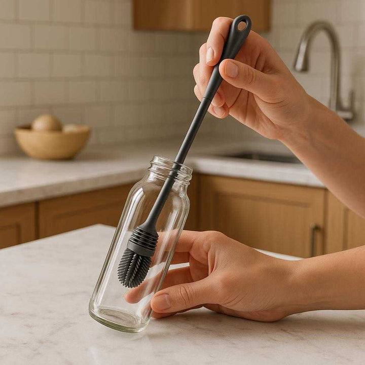 Boremaskine Rengøringsbørste til Flasker i Grå Silikone og Plast: A realistic kitchen setting where the product is properly grounded on a marble countertop, being actively used by a person's hand to clean the inside of a glass bottle.