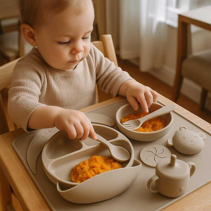 Børneservice i Silikone - Professionelt Spisesæt med Sugekop, Bestik og Bidering i Beige: A realistic family dining room setting where the silicone eating set is properly grounded on a wooden high chair tray, being actively used by a toddler.