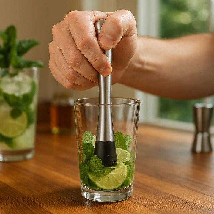 Cocktailpind i Rustfrit Stål - Genanvendelig og Stilfuld til Drinks og Appetizers, Tilgængelig i Sort og Sølv: A realistic home bar setting where the cocktail muddler is properly grounded on a wooden counter, being actively used by a person's hand to crush mint leaves and lime slices in a glass for a mojito.
