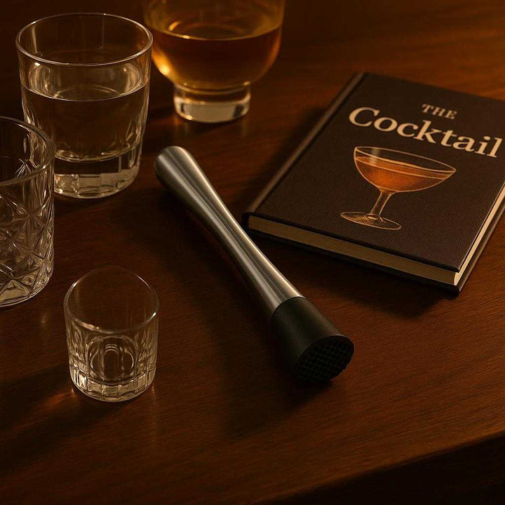 Cocktailstøder i Rustfrit Stål med Rillet Bund - Professionel Høj Kvalitet til Hjemmebaren. The elegant stainless steel cocktail muddler rests next to a stylish home bar setup, complete with glasses and a cocktail recipe book, under warm ambient lighting.