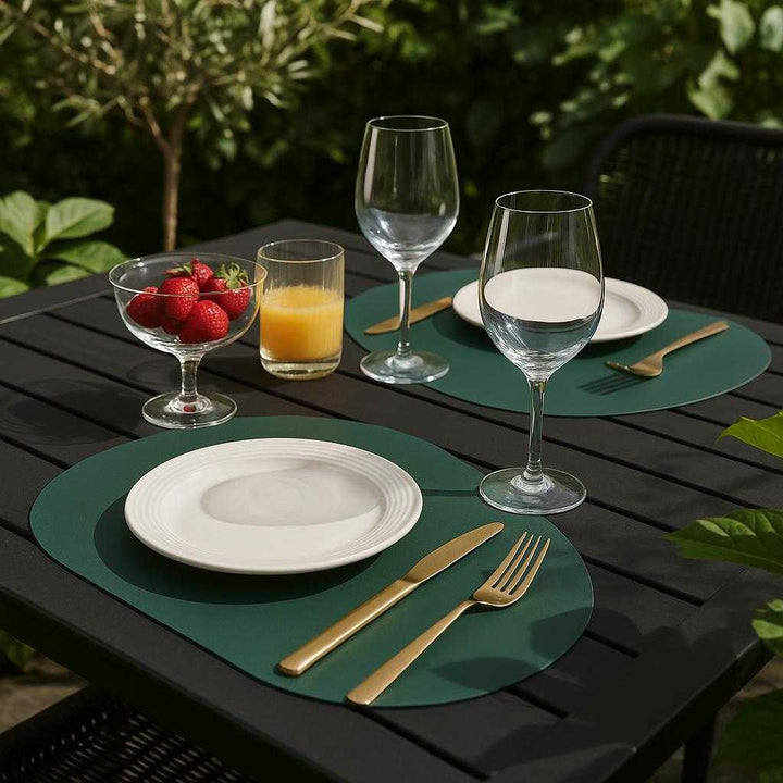 Dækkeserviet i mørkegrønt PU-læder - Sæt med 8 stk. i moderne design. The dækkeserviet is prominently placed on a stylish outdoor patio table, surrounded by lush greenery and a set of chic tableware, capturing the essence of a sophisticated summer brunch with friends.