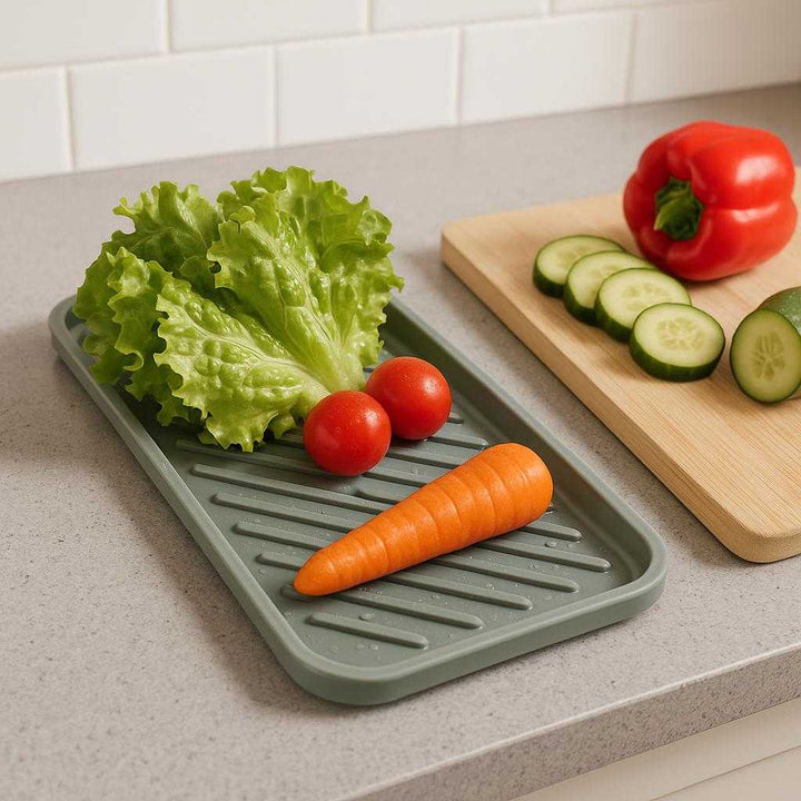 Drypbakker i Silikone til Grill og Husholdningsbrug - Grå, Hvid, Orange, Sort - Høj Kvalitet og Funktionalitet. The versatile silicone drip tray is neatly positioned on a modern kitchen countertop, catching water and debris from freshly washed vegetables beside a cutting board with colorful produce.