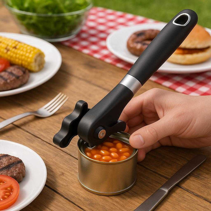 Dåseåbner i rustfrit stål - Ergonomisk design, nem at bruge og effektiv til åbning af dåser: A different realistic picnic table setting where the can opener is securely positioned on a wooden table, being used to open a can of beans for a barbeque, with proper physics, shadows, and realistic integration into the environment, surrounded by picnic items like plates and cutlery.