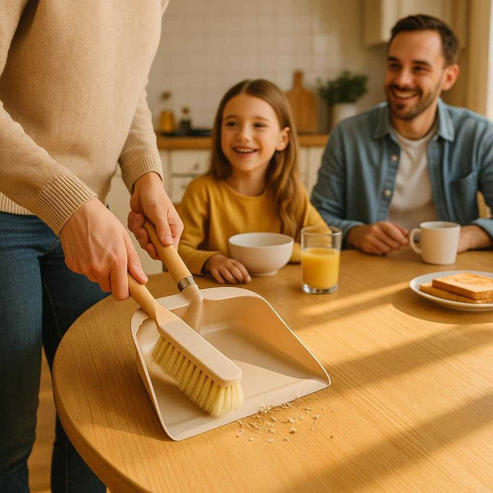 Fejebakke og kost i moderne design - Kompakt og nem at opbevare, i beige og træ. In a sunlit kitchen, the beige dustpan and brush set is skillfully used to sweep crumbs from a dining table, with a cheerful family enjoying breakfast nearby, emphasizing its practicality and modern design.