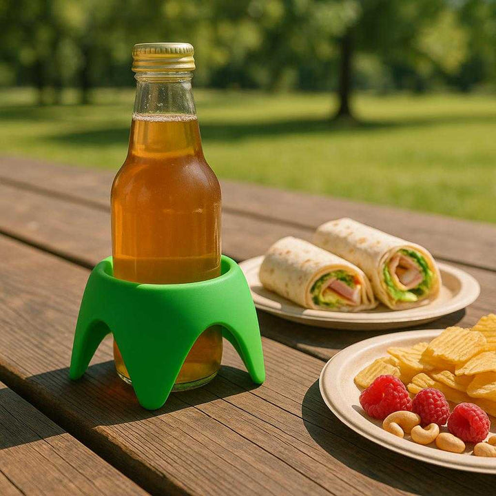 Flaskeholder i Silikone - Praktisk Støtte til Flasker i Blå, Grøn, Lilla og Orange: A different realistic outdoor picnic setting where the silicone bottle holder in green is securely positioned on a wooden picnic table, holding a beverage bottle alongside a spread of sandwich wraps and snacks, with proper physics, shadows, and integration into the sunny, park-like environment.