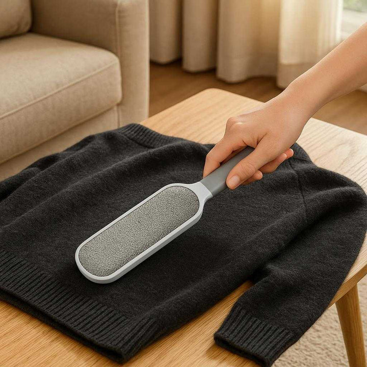 Fnugfjerner i håndholdt design - Effektiv fjernelse af fnug og fibre fra tekstiler - Grå: A realistic living room setting where the lint remover is properly grounded on a coffee table, being actively used by a hand to clean a dark woolen sweater draped over the table.