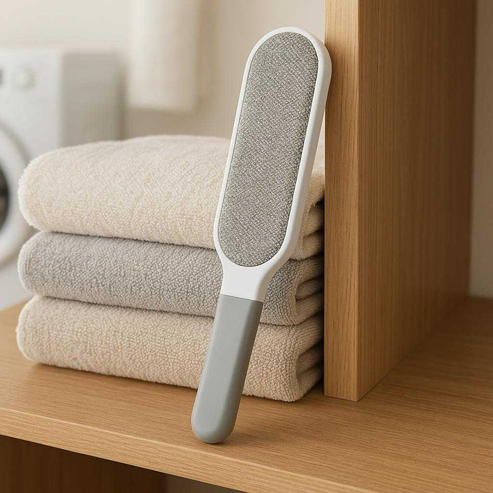 Fnugfjerner i håndholdt design - Effektiv fjernelse af fnug og fibre fra tekstiler - Grå: A different realistic laundry room setting where the lint remover is securely positioned on a wooden shelf, naturally placed among folded clothes.