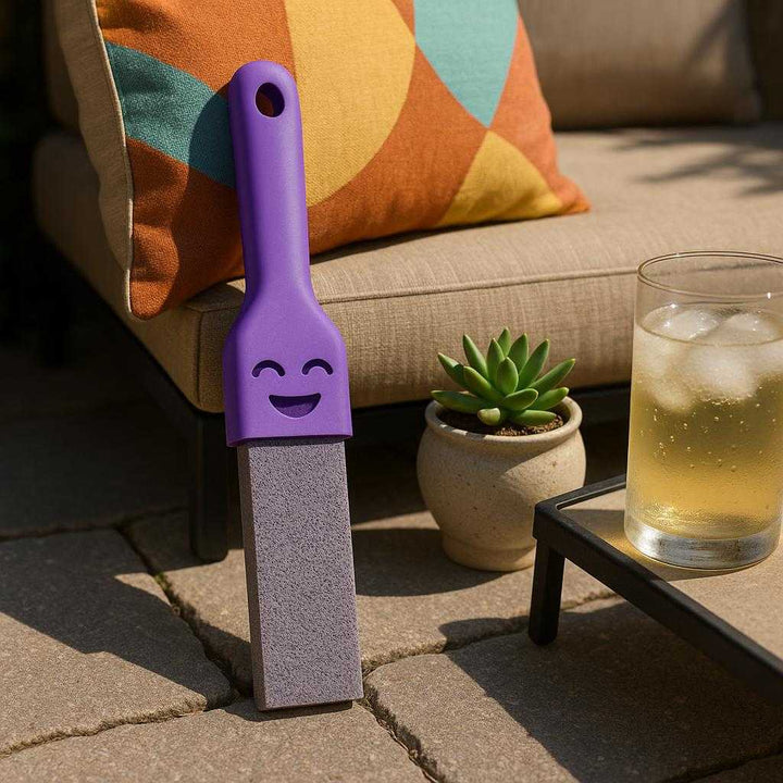 Fugerenser med effektiv slibesten og ergonomisk greb – Tilgængelig i Lyserød, Lilla og Gul. An outdoor patio with stone flooring, where the product is used to clean between the stone joints. The purple-handled version rests against a colorful outdoor cushion, next to a small ceramic planter. Sunlight casts soft shadows, illuminating the rugged effectiveness of the slibesten. A cold drink with condensation sits on a table, enhancing the feeling of a relaxed, well-maintained space.