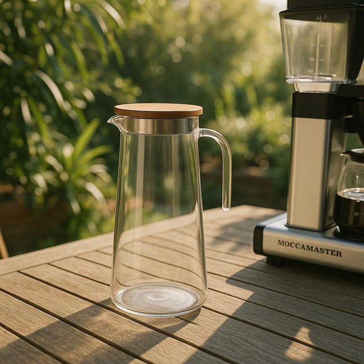 Glaskande med Låg og Hældetud i Træ – Høj Kvalitet og Elegant Design til Vand og Moccamaster. The stylish glass carafe sits on an outdoor patio table, casting a soft shadow, beside a Moccamaster coffee maker, with morning sunlight streaming through lush garden plants in the background.
