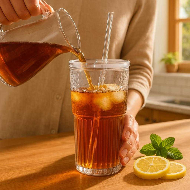Glaskop med låg og sugerør - Praktisk og funktionel til kolde drikke: A realistic sunny kitchen setting where the glass tumbler with lid and straw is properly grounded on a wooden kitchen counter, being actively used by a person.
