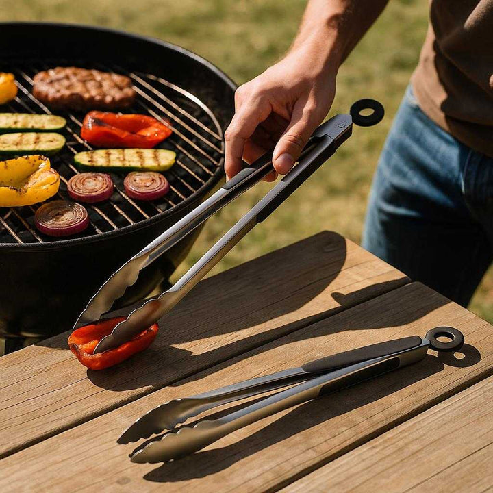 Grilltang i Rustfrit Stål med Låsemekanisme - Professionel og Sikker Madlavning: A realistic outdoor barbecue setting where the stainless steel grill tongs are properly grounded on a wooden picnic table, actively used by a person flipping grilled vegetables and meat over a hot grill, with natural sunlight casting realistic shadows and showing clear contact points on the table surface.