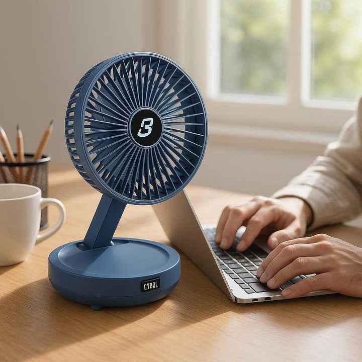 Håndventilator med Batteri - Moderne, Støjsvag og Foldbar Design i Blå og Hvid Farve: A realistic office desk setting where the fan is properly grounded on a wooden desk, slightly angled upward, being actively used by an individual working on a laptop.