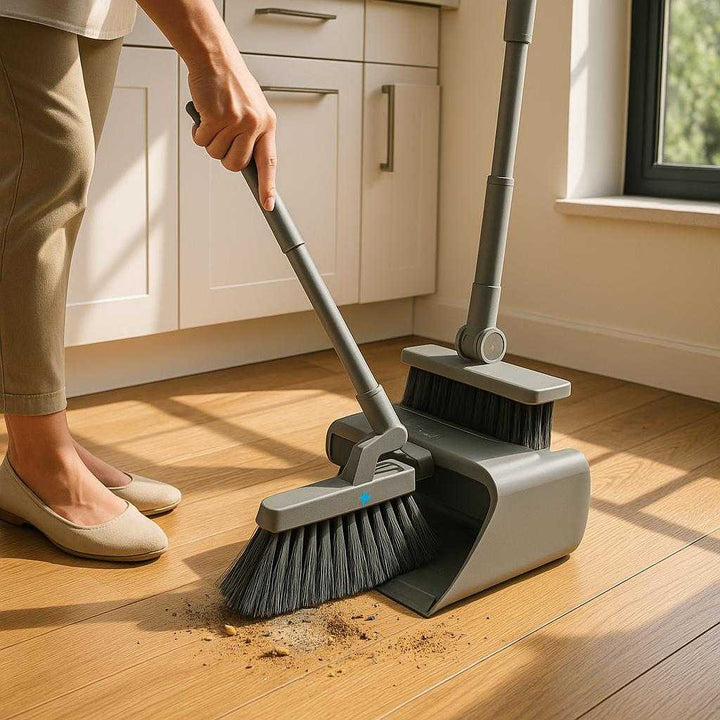Integreret Fejesæt med Kost, Skraber og Opbevaring i Grå og Hvid - Effektiv Rengøring med Stil. The integrated gray and white cleaning set with broom and scraper is actively being used to sweep dust and debris off a hardwood floor in a modern kitchen, sunlight streaming through a nearby window.