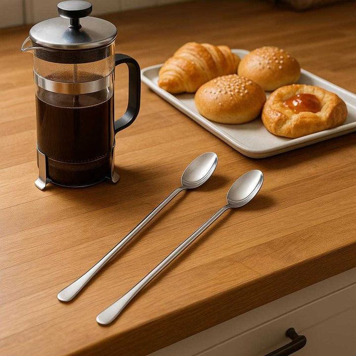 Iskaffeske i rustfrit stål – Lang, Elegant og Funktionel til Høje Glas, 2 stk.. The stainless steel iced coffee spoons elegantly resting on a wooden kitchen countertop beside a French press and a tray of assorted pastries, evoking a sophisticated breakfast setting.
