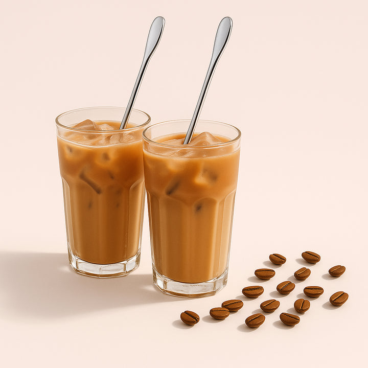 Enhanced Iskaffeske i rustfrit stål – Lang, Elegant og Funktionel til Høje Glas, 2 stk.. Two elegant stainless steel iced coffee spoons stirring creamy iced lattes in tall clear glasses on a sunlit cafe table, with coffee beans and a small potted plant nearby.