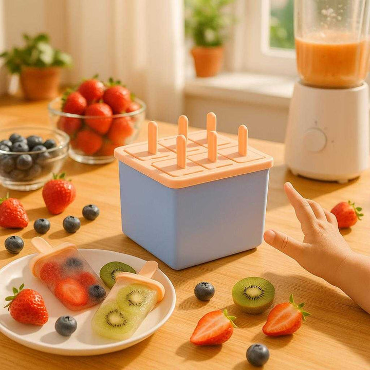 Ispindeform i moderne design - Lav dine egne ispinde nemt og hurtigt - 6 rum - Blå og Rosa. A bright, sunny kitchen with natural light streaming through the windows, where the ice mold is sitting on a wooden countertop filled with colorful fruit ingredients. A couple of half-prepared popsicles are visible with fresh berries and kiwi nearby. A child’s hand reaches towards the mold, expressing excitement and anticipation, while a blender with fruit puree is seen in the background.