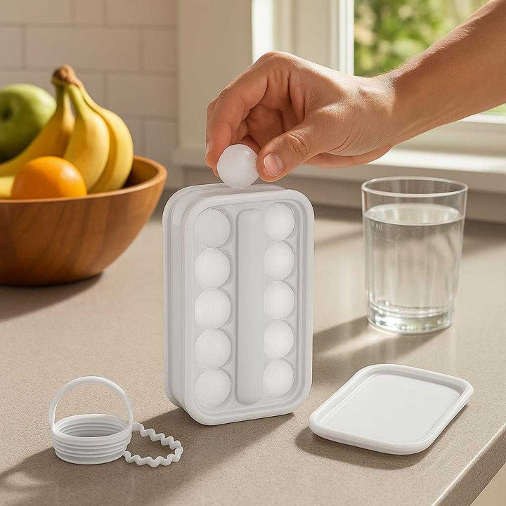 Isterningbakke med låg og skubber – Fleksibel silikoneform til nem udtagning, perfekt til både hverdag og fest: A realistic kitchen setting where the ice tray is properly grounded on a sleek kitchen counter beside a fruit bowl, its lid off exposing the ice balls inside, being actively used by a hand taking out ice for a refreshing drink.