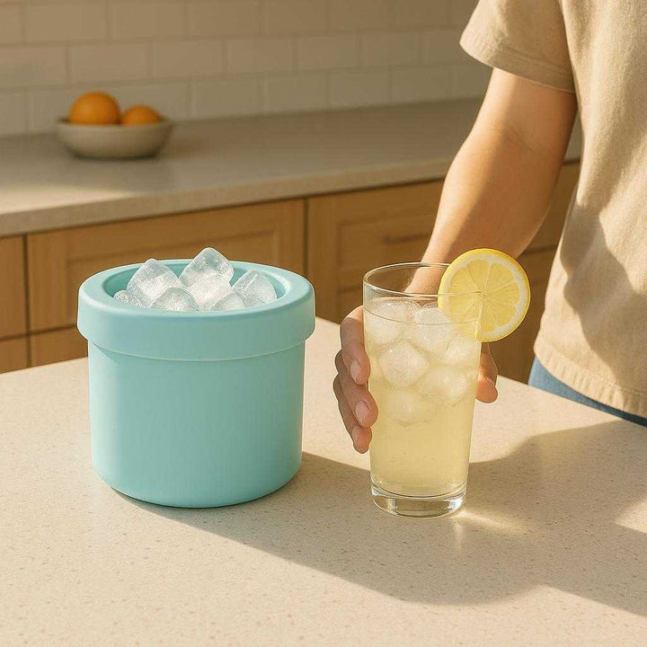 Isterningebøtte i Silikone – Praktisk og Kompakt Opbevaring i Blå, Grå og Gul. The silicone ice bucket in blue is perched on a kitchen counter, filled with ice cubes, while a person reaches for an iced lemon drink on a sunny afternoon.