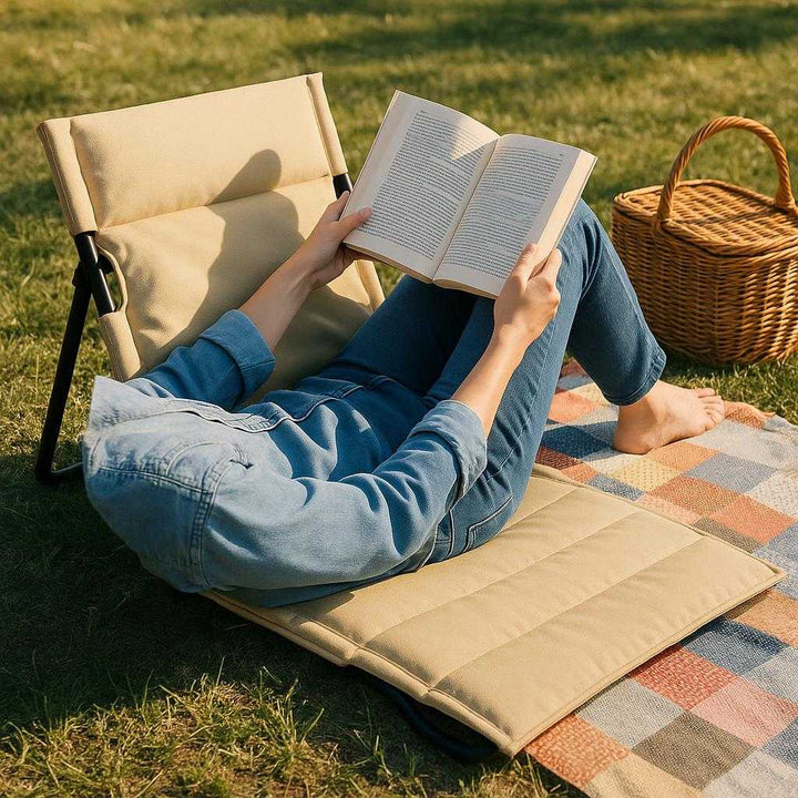 Klap stol i beige, brun, grøn, sort - Stof, metal, polstret sæde - Bærbar og komfortabel: A realistic outdoor picnic setting where the folding chair is properly grounded on a grassy field, being actively used by a person relaxing while reading a book.