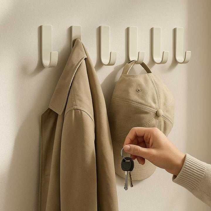 Knagerække i Hvid Mat Metal - Selvklæbende og Pladsbesparende til Hjemmet: A realistic entryway setting where the product is properly grounded on a smooth, painted wall, being used to hang a coat, hat, and keys by a person.