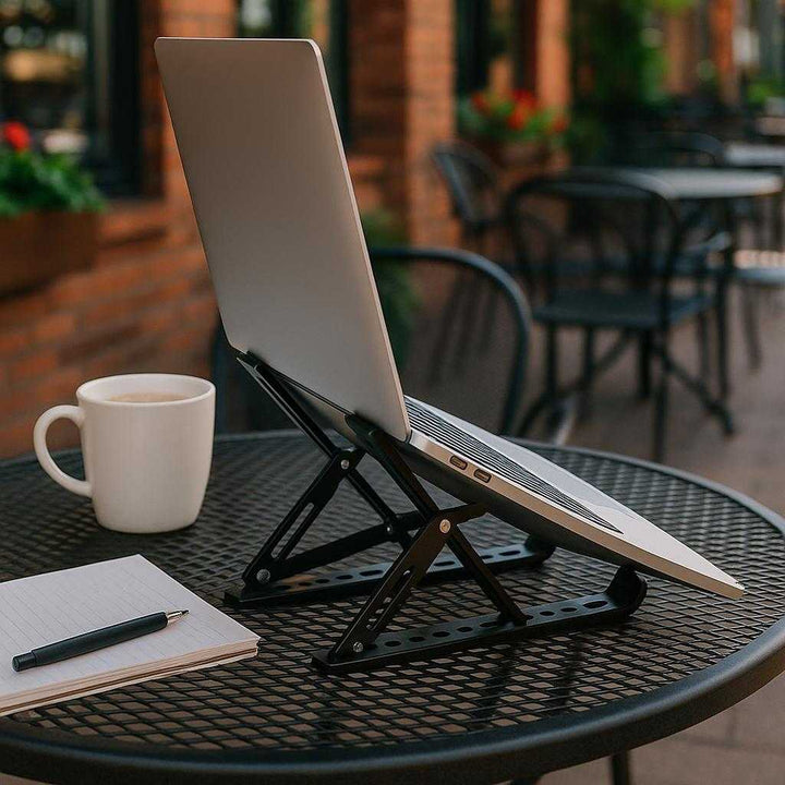 Laptop stand i sort og hvid - Justerbar og foldbar til bedre ergonomi og luftcirkulation: A different realistic cafe environment where the laptop stand is securely positioned on a metal outdoor table, supporting a laptop naturally placed for use.
