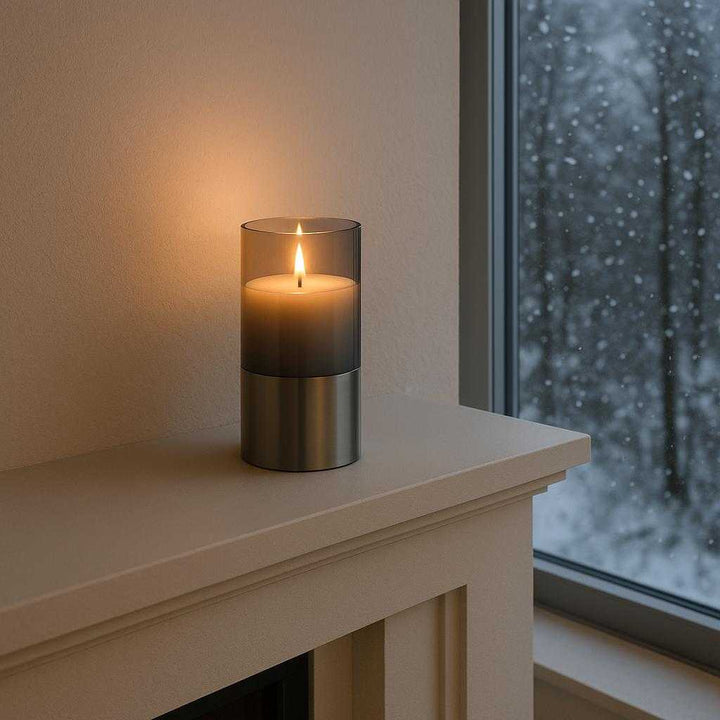Ledlys med autentisk flammeeffekt - Moderne og minimalistisk design i sølv og grå. The candle is nestled on a minimalist mantelpiece in a modern living room, casting a cozy light as gentle snow falls outside a large window, enhancing the room's serene and peaceful ambiance.