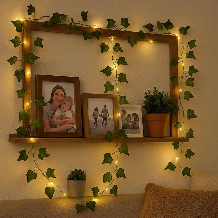 Lyskæde med Kunstige Blade - Dekorativ Belysning til Indendørs Brug, Energibesparende LED, Grøn & Varm Hvid: A cozy living room setting where the decorative light string with artificial leaves is draped along a wooden shelf, surrounded by framed family photos and small potted plants, casting warm, inviting glows on the nearby wall.