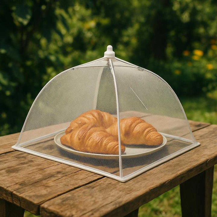 Madkuppel i Finmasket Net - Elegant Beskyttelse til Dine Retter med Stil og Funktionalitet. The elegant mesh food cover is placed over a plate of freshly baked croissants on a rustic outdoor wooden table, surrounded by a lush garden setting.