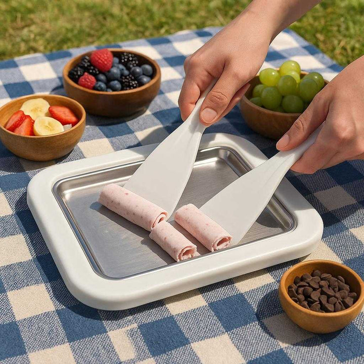 Maskine til Hjemmelavede Isruller - Ingen Strøm Nødvendig, Perfekt til Børn og Dessertelskere: A different realistic outdoor picnic setting where the ice roll maker is securely positioned on a picnic blanket, surrounded by bowls of fruit and toppings.