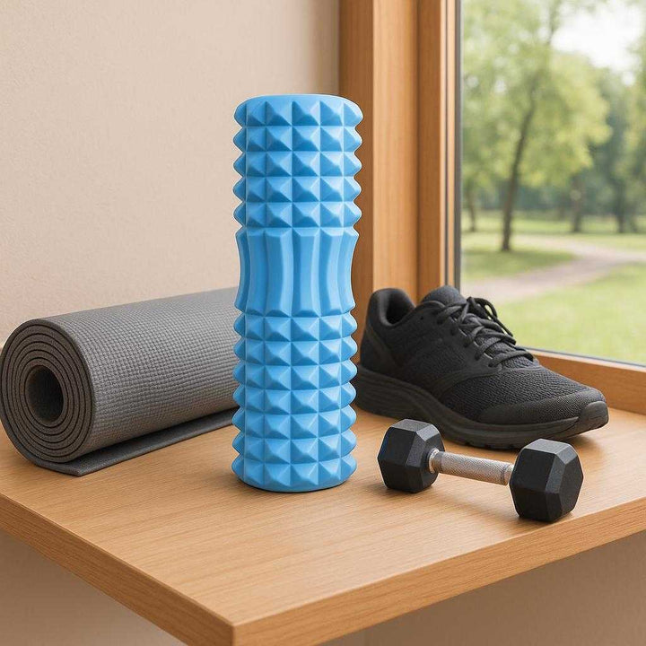 Massagerulle med Dybdegående Struktur i Blå, Lilla, Lyserød, Sort – Professionel Restitution & Komfort. The blue massage roller rests on a wooden shelf beside gym gear, with a window overlooking a scenic park view in the background, evoking a sense of relaxation and wellness.