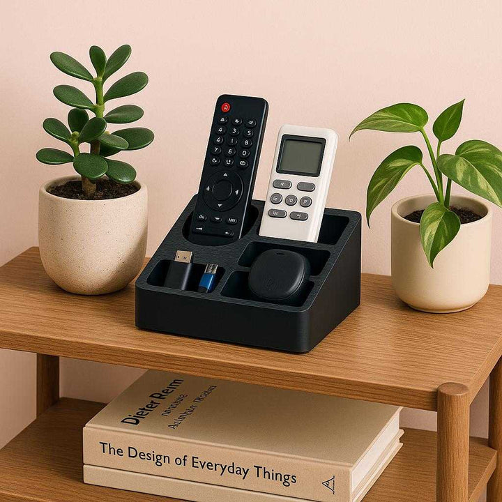 Opbevaringsholder i Sort Plast med Flerdelt Ruminddeling - Professionel Høj Kvalitet til Kontor og Hjem. A stylish home office setting featuring the black plastic organizer neatly storing remote controls and small gadgets on a wooden shelf, accompanied by decorative plants and books.