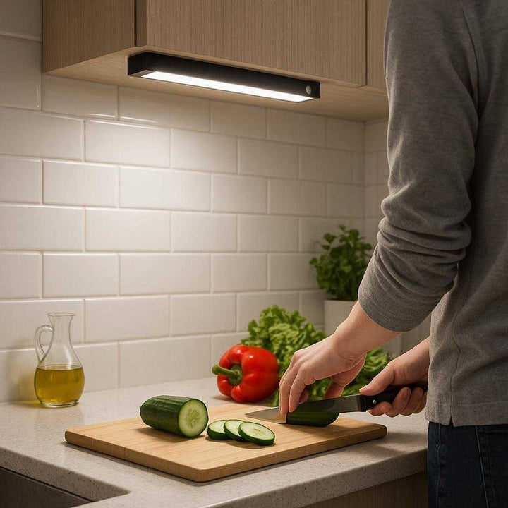 Opladeligt LED-skabslys med bevægelsessensor - Moderne og energieffektiv belysning til hjemmet i sort og lyseblå: A realistic kitchen setting where the product is properly grounded under a cabinet, illuminating a countertop as a person prepares food nearby, with natural lighting casting realistic shadows and showing clear contact points where the product touches the cabinet surface.