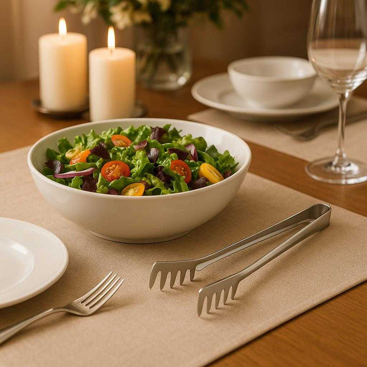Pastatang i Rustfrit Stål - Professionel Kvalitet med Silikonegreb til Pasta og Salater. The elegant stainless steel pasta tongs are placed gracefully on a dining table next to a vibrant salad bowl, highlighting a sophisticated dinner setting with candles and fine china.