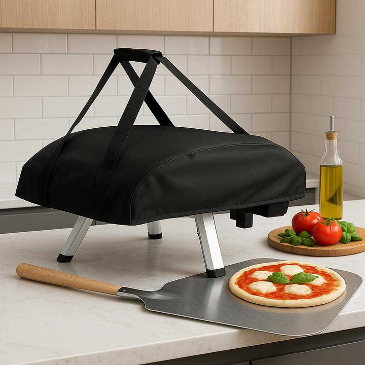 Pizzaovn Bærbar Gas og Elektrisk – Perfekt til Sprøde Hjemmelavede Pizzaer i Høj Kvalitet. The sleek, covered portable pizza oven sits atop a modern kitchen counter, ready to be carried outdoors, with a pizza peel and fresh ingredients nearby.