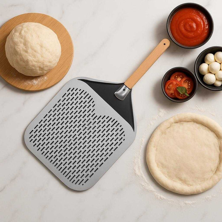 Pizzaspade med huller - Professionel og letvægts, tilgængelig i sølv og sort. The sleek silver and black pizza peel rests on a modern kitchen countertop, next to freshly prepared pizza dough and toppings, evoking a gourmet cooking atmosphere.