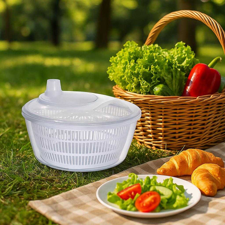 Salatslynge med håndsving og si - Effektiv tørring og servering af salat og grøntsager: A different realistic picnic setting outdoors where the product is securely positioned on a grassy surface, naturally placed next to a picnic basket filled with fresh vegetables.