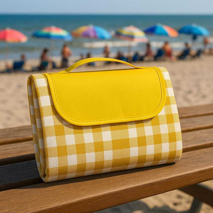 Sammenfoldeligt Picnictæppe med Bærehåndtag i Gul og Rød - Høj Kvalitet og Funktionelt Design. The folded picnic blanket with its convenient carry handle is perched on a wooden bench at a bustling beach with colorful umbrellas in the distance.