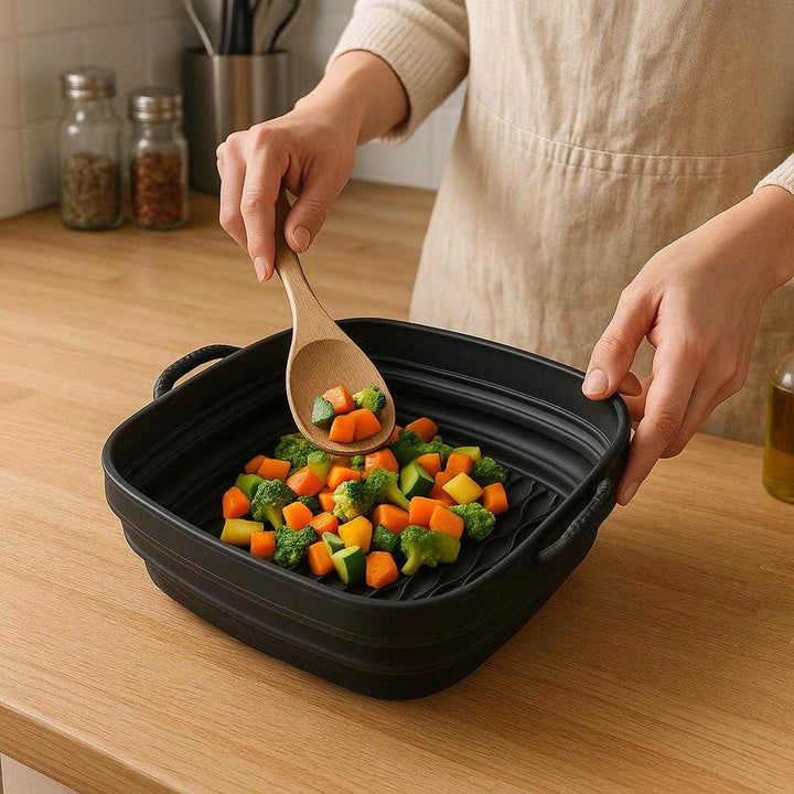 Sammenklappelig Airfryer Silikoneform Firkantet - Effektiv Madlavning, Fleksibel Design, Høj Kvalitet: A realistic kitchen setting where the Sammenklappelig Airfryer Silikoneform Firkantet is properly grounded on a wooden countertop, being actively used by a person placing chopped vegetables inside with a spatula in another hand.